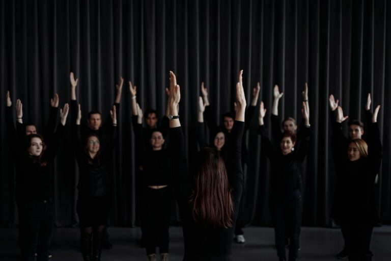 A group of performers rehearsing a contemporary dance with raised arms indoors.